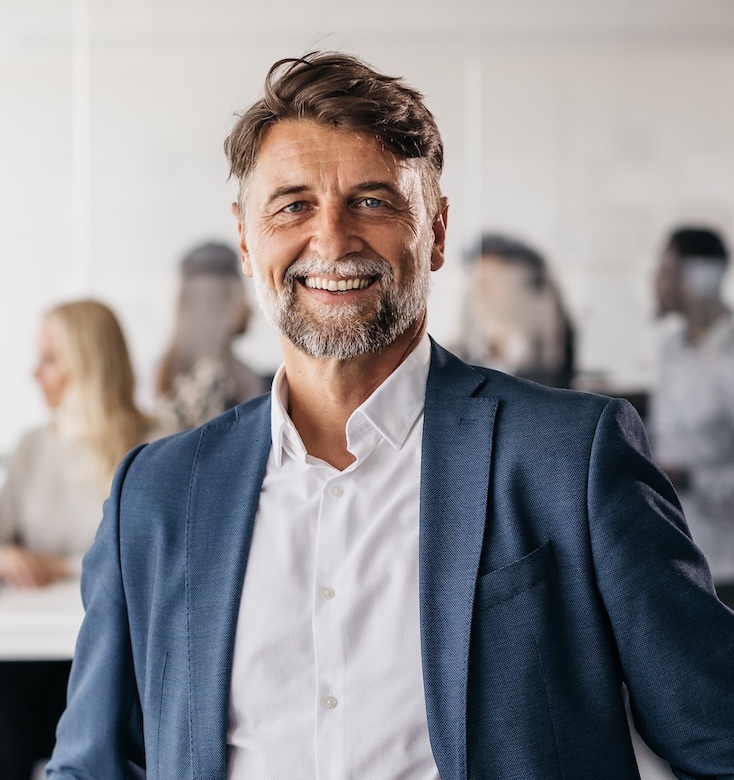 A successful businessman is standing in a spacious office and smiling at the camera. He is dressed in a fashionable business suit. His colleagues are working in the background. Horizontal daylight indoor photo.